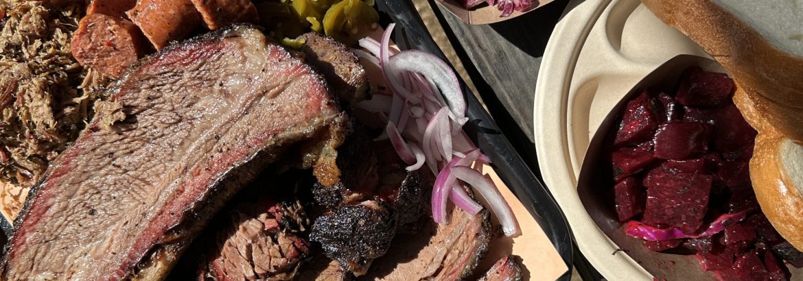 An overhead view of a large tray of Texas barbecue foods, including beef rib, beef brisket, sausage, pulled lamb, plus sides of pickles, mac and cheese, grits, beets, bread, and more.
