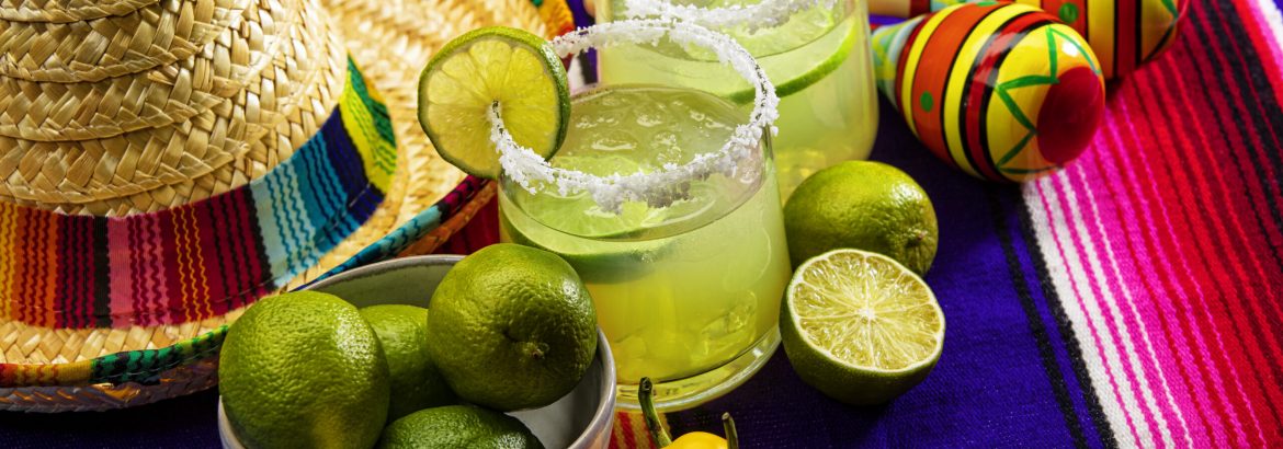 This is a photograph of two modern margarita glass with a rim of salt surrounded by fresh cut limes and chilis on a colorful striped mexican blanket next to a sombrero