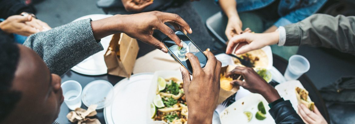 A multi-ethnic group of friends enjoy tacos and chips at a Mexican food cart in downtown New York City, New York.  They laugh and talk while eating their food, having fun sharing life and culture.  One of them takes a picture of their food with his smartphone to share on his social networks.  Horizontal image.