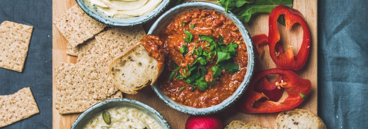 Vegan snack board. Flat-lay of Various Vegetarian dips hummus, babaganush and muhammara with crackers, bread, fresh vegetables on wooden board over grey background. Clean eating, dieting food concept