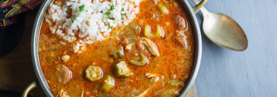 Top view from directly above of a small copper pot of chicken and sausage gumbo made with fresh okra and tomatoes. Gray background on cutting board.