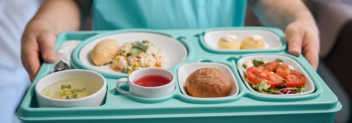 Cropped photo of nursing assistant holding meal tray in hands while standing in hospital room
