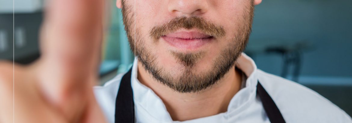 Confident mid adult Hispanic personal chef selects items for a salad from digital image. He is wearing chef's whites and a black apron. He has brown hair and facial hair.