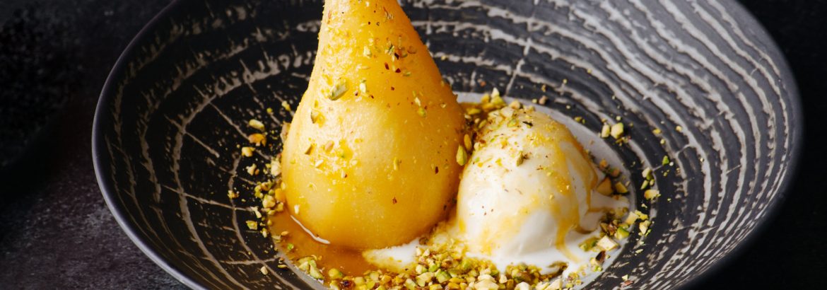 Boiled pear and icecream sprinkled with nuts in exquisite dessert. On a black plate on a black table.