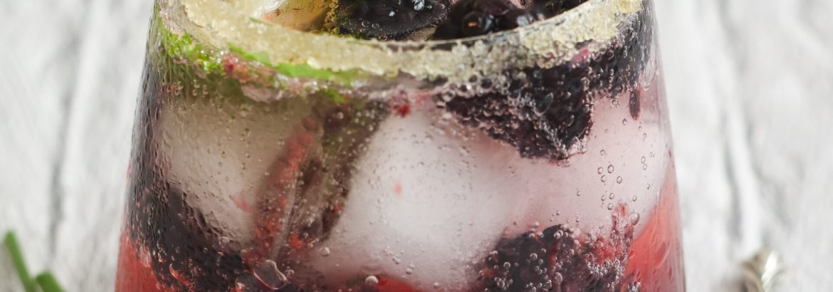 A glass of iced colorful blackberries citrus margarita non-alcohol with sugar at the edge of glass. With its fruits in spoon and purple flowers in background. A ideal fresh cold summer drink
