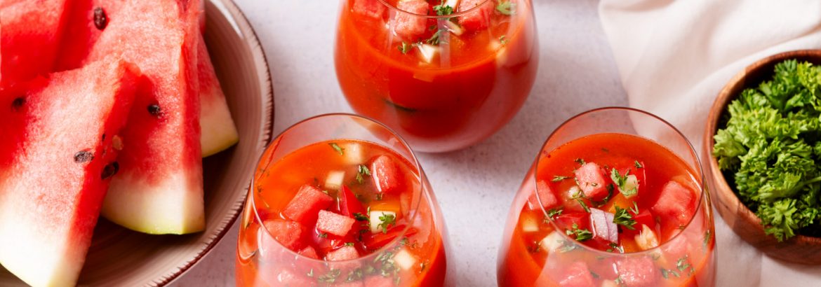 Spanish tomato and watermelon gazpacho cold soup styled and decorated in glasses with ingredients on white table, top view