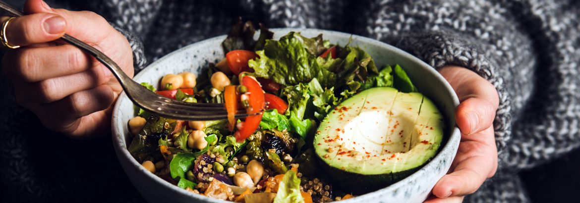 Healthy vegetarian dinner. Woman in grey jeans and sweater eating fresh salad, avocado half, grains, beans, roasted vegetables from Buddha bowl. Superfood, clean eating, dieting food concept