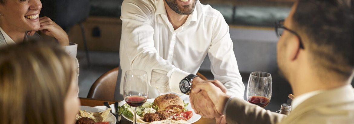 Happy businessmen shaking hands while having a lunch with their females colleagues in a restaurant.